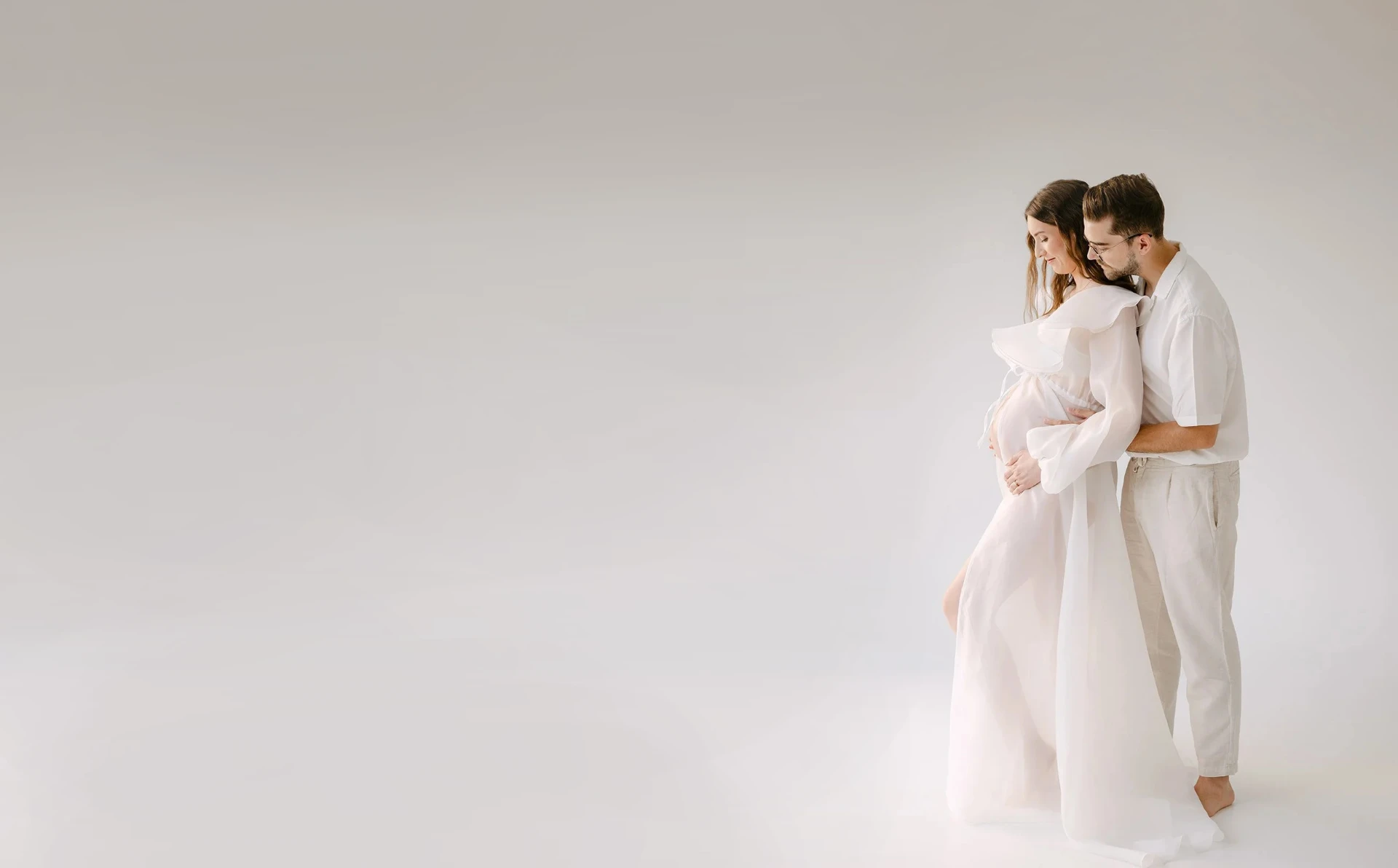 Expecting couple in soft white clothing embracing in a light studio setting